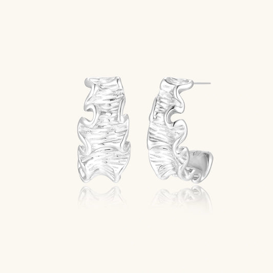 Textured white gold-plated hoop earrings with flowing wave pattern against cream background, showing dimensional rippled surface.