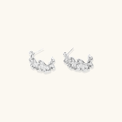 Textured white gold-plated hoop earrings with flowing wave pattern against cream background, showing dimensional rippled surface.